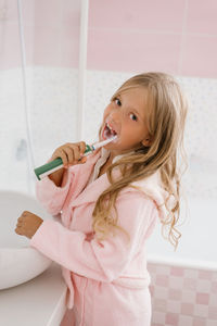 Cute child girl in a pink terry dressing gown brushes her teeth in front of the bathroom mirror