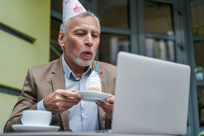 Smiling senior man wearing party hat holding cake video calling while sitting at cafe