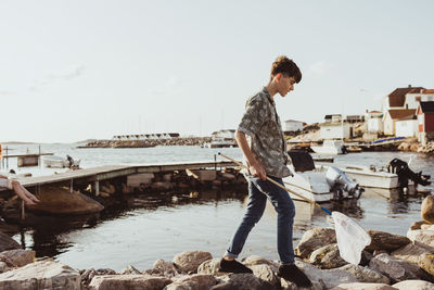 Teenage boy with fishing net walking over rocks against sky