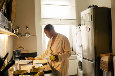 Young woman holding grapes using laptop in kitchen at home