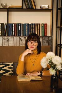 Portrait of young woman reading book on table in library