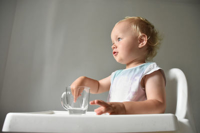 Almost 2 year old baby drinks water from transparent glass, side view