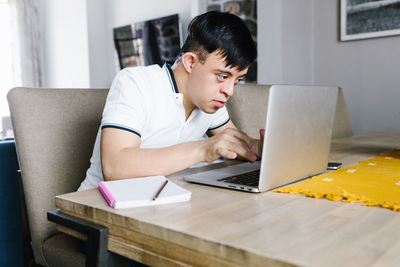 Focus latin teen boy with down syndrome browsing netbook while sitting at table and studying online from home