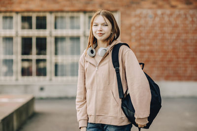 Portrait of smiling teenage female student with backpack and headphones standing in high school campus