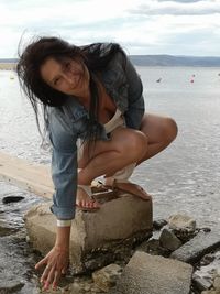 Portrait of woman crouching at beach against sky