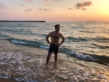 Shirtless man standing on beach against sky during sunset