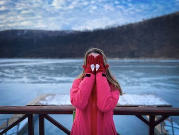 Young woman standing near a frozen lake and covering her face with her hands wearing red gloves