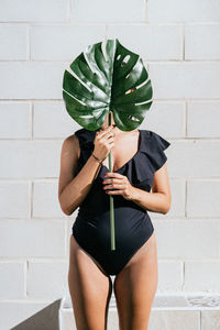 Unrecognizable female in stylish black swimwear hiding face behind green monstera leaf while resting on poolside near wall in sunny summer day