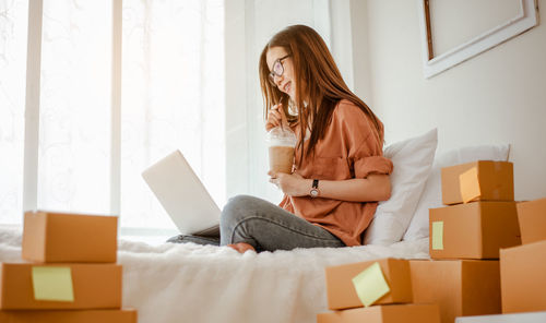 Side view of young woman with coffee using laptop on bed in home office