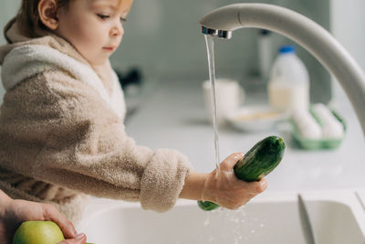 A little girl washes cucumbers in the sink in the kitchen.