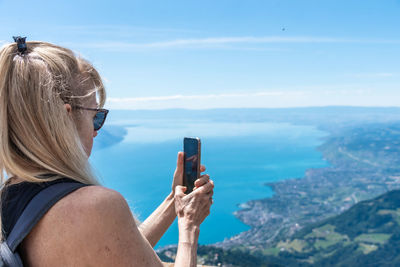 Mature blonde woman with sunglasses and backpack taking a photo of lake geneva from a mountain.