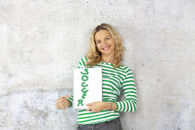 Portrait of smiling beautiful woman holding book with text while standing against wall