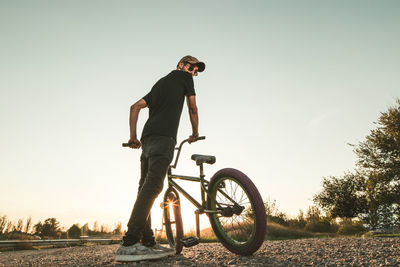 Low angle view of young man with bicycle standing on field against clear sky during sunset
