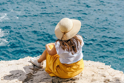 Young woman in summer clothes and hat sitting on edge of cliff above sea.