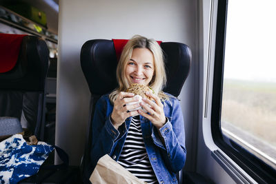 Portrait of smiling blond woman travelling by train having a snack