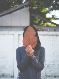 Woman covering face with dried leaf while standing against surrounding wall