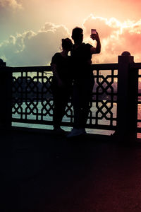 Silhouette man photographing on railing against sky during sunset