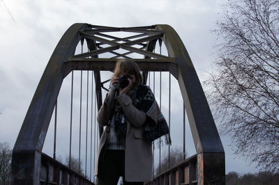 Mature woman photographing while standing on bridge against cloudy sky