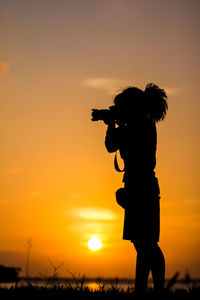 Silhouette woman photographing using camera against orange sky during sunset