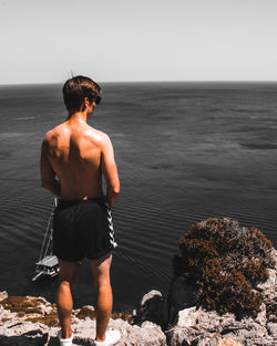 Rear view of shirtless man looking at sea while standing on cliff against sky