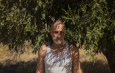 Adult man in white tank top and jeans standing with olive tree in summer