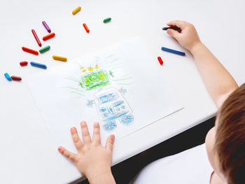 Toddler draws robot. kid uses wax crayons. top view on child hands and pencils on white background.