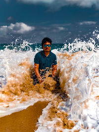 Portrait of man wearing sunglasses while sitting at beach with splashing water