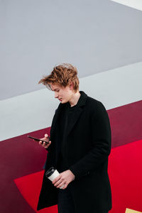 Young man in black coat using smartphone with cup of coffee in hands looking away outdoors