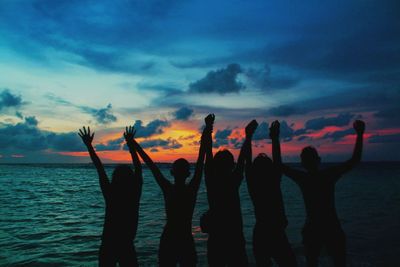 Silhouette people with arms raised standing in sea during sunset