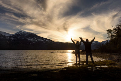 Friends standing by lake against sky during sunset