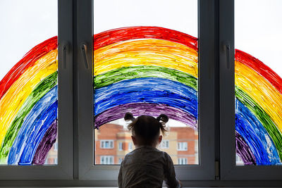 Little girl on background of painting rainbow on window. kids leisure at home. positive visual