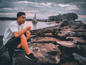 Young man sitting on rock by sea against sky