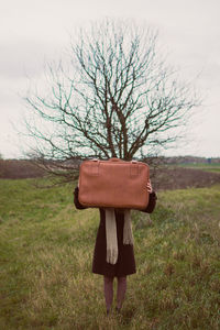 Woman covering face with briefcase on field against sky