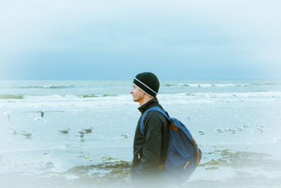 Side view of thoughtful man with backpack standing on sea shore at beach