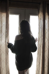 Rear view of woman standing against window at home