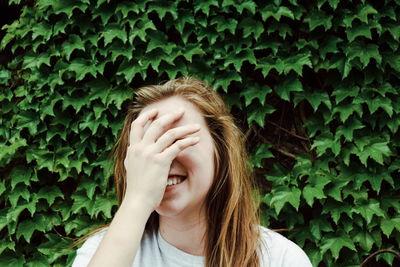 Smiling young woman covering face while standing against plants