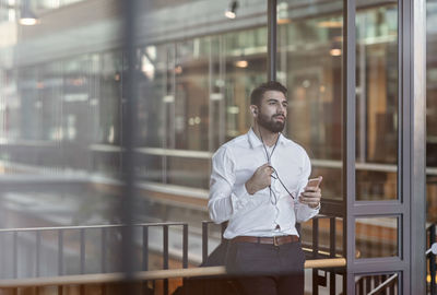 Businessman wearing headphones and using smart phone while standing in lobby at office