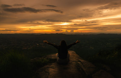 Rear view of woman sitting on cliff against sky during sunset