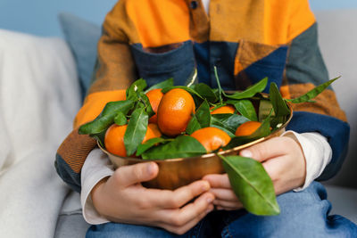 Boy in a bright blue-orange checkered shirt and jeans holds a copper bowl of fresh tangerines.
