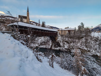 Snow covered buildings and bridge by trees against sky