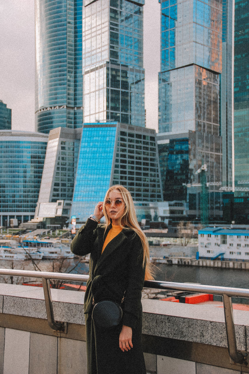 city, one person, architecture, building exterior, standing, hair, built structure, city life, young adult, office building exterior, adult, women, smiling, long hair, skyscraper, building, young women, hairstyle, blond hair, beautiful woman, modern, outdoors, cityscape