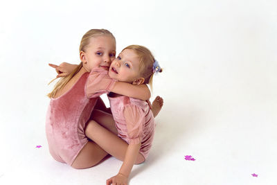 Two sisters sitting on a white background in pink bodysuits