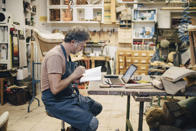 Side view of male upholstery worker reading book while sitting at workbench in workshop