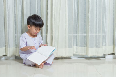 Boy reading book while sitting against curtain at home