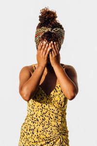 Smiling african american female wearing summer dress and trendy headband standing near wall in city and covering face while looking at camera