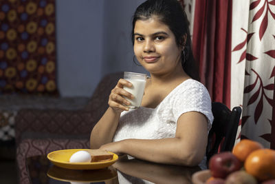 An indian asian woman holding a glass of milk sitting beside a table in a domestic room