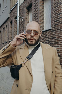 Portrait of man talking on phone while standing against building on footpath