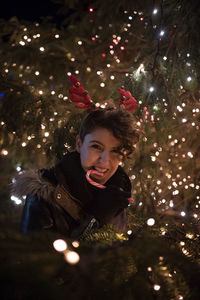 Happy young woman eating candy cane by illuminated christmas tree at night