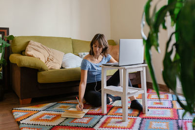 Focused adult female remote employee sitting on floor in front of laptop and taking notes in planner while working online at home