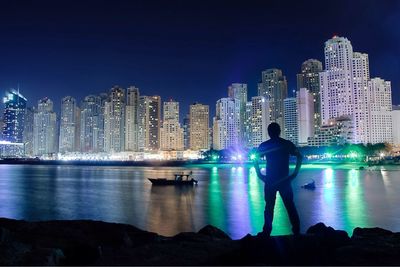 Silhouette man standing at lakeshore against illuminated cityscape